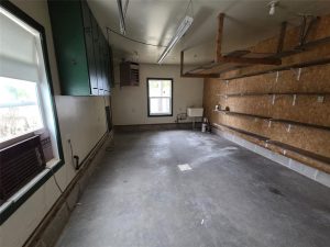 A mostly empty garage with concrete floors, wall shelves on the right, green cabinets on the left, a window with an AC unit, a sink at the back, and overhead fluorescent lights.