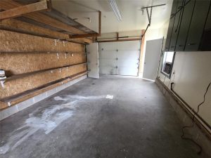 Empty garage with concrete floor, wooden shelves on the left wall, closed overhead garage door at the back, open side door, green cabinets on the right wall, and fluorescent lighting on the ceiling.