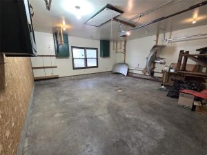 An empty garage with a concrete floor, shelves, green cabinets, a window, auto parts, black trash bags, and a box stacked against the walls. Overhead lights illuminate the clean, spacious area.