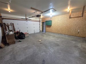 A clean, empty garage with a concrete floor, a closed white double garage door, plywood walls, a few shelves, cleaning supplies, a broom, boxes, and a green electrical panel on the back wall.