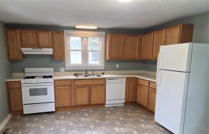 A kitchen with light wood cabinets, white appliances including a stove, range hood, dishwasher, and refrigerator, a double sink under a window, and gray walls with a patterned linoleum floor.