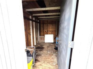 View inside a small, empty wooden shed with exposed beams and plywood walls. The door is open, revealing a few scattered items, including a yellow bin and a white object leaning against the wall.