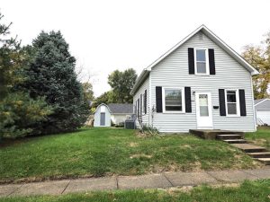 A small white house with black shutters and a gabled roof sits beside a grassy yard and a large pine tree. Concrete steps lead up from the sidewalk. A small shed is visible in the background.