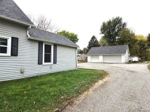 A gravel driveway curves past a gray house with black shutters and leads to a large detached garage; green grass and trees with autumn foliage surround the buildings under a cloudy sky.