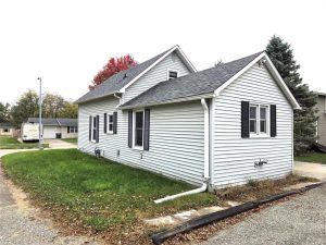A small white house with black shutters, a gray shingled roof, and green grass lawn sits on a corner lot in a suburban neighborhood. Trees and other houses are visible in the background.