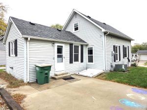 A small, light gray house with white trim and a dark shingled roof, featuring a side entrance, green trash bin, concrete driveway, grassy yard, and wooden access ramp. The scene appears to be in a residential neighborhood.
