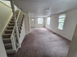 A mostly empty living room with carpeted floors, white walls, two windows, a white front door, and a staircase with a white railing leading upstairs. Natural light comes through the windows.