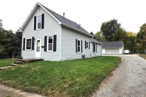 A white two-story house with black shutters and a gray roof sits on a green lawn at 305 N 4th Street, Indianola, Iowa. A gravel driveway curves beside the home, leading to a detached garage surrounded by trees—a prime real estate opportunity.