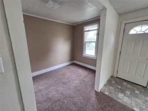 Small room with beige walls, white trim, and brown carpet next to an entryway with a white door and tiled floor. A window lets in natural light. Ceiling is white with square patterns.