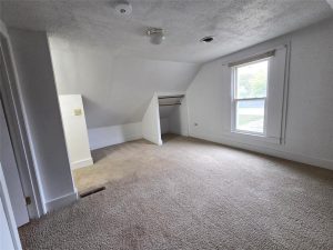 An empty, carpeted room with white walls, a large window with blinds, a small open closet with a wooden rod, and a sloped ceiling. Natural light enters through the window, brightening the space.
