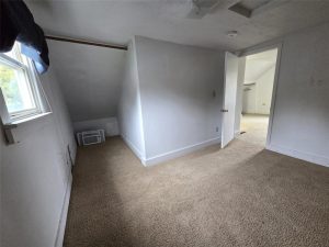 A small, empty room with beige carpet, white walls, a slanted ceiling, a single window with a blue curtain, and an open door leading to another room. An air vent is on the floor near the corner.