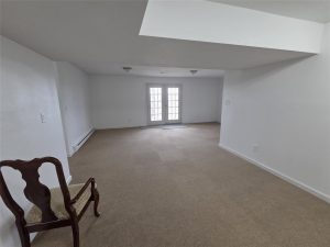 A mostly empty room with beige carpet, white walls, a single wooden chair, and double glass doors at the far end letting in light. Ceiling has an angled drop section.