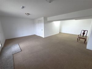 Spacious, empty room with white walls, beige carpet, and two ceiling lights. A single wooden chair is placed in one corner near a door. A small area of the carpet near a window appears to be a different shade.