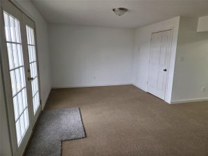 A mostly empty room with beige carpet, a small gray rug, white walls, a ceiling light, French doors on the left, and a closed white door on the right wall.