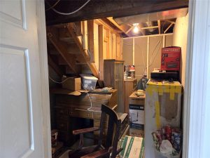 A cluttered basement storage area with exposed ceilings, wooden stairs, old furniture, a Craftsman toolbox, cardboard boxes, and various household items scattered around under a single hanging light bulb.