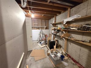A cluttered basement or utility room with exposed ceiling beams, unfinished walls, shelves filled with tools and supplies, and a ladder near a closed white door at the far end. Construction materials are scattered on the floor.