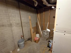 A basement room with unfinished walls, exposed pipes, a bucket, a cardboard box with pink tape, wooden planks, a floor fan, and scattered construction materials on a concrete floor.