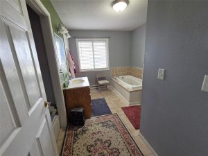 A bathroom with gray walls, a white ceiling, and tile floors features a large rug, a wooden vanity with a sink, a mirror, a window with blinds, a folding chair, and a corner bathtub with tan tile surround.