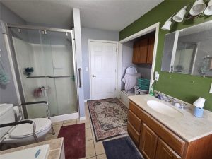 A bathroom with a walk-in shower, toilet with grab bars, wooden vanity, mirror, washing machine, laundry baskets, and cabinets. The walls are green and gray, with tiled flooring and an area rug.