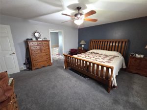 A bedroom with gray carpet, a wooden bed with a patterned quilt, matching wooden dressers and nightstands, a ceiling fan, and an open doorway leading to a bathroom.