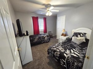 A bedroom with two twin beds covered in black and white floral bedding, a TV on a white dresser, a rocking chair by the window with red curtains, and a ceiling fan with light. The walls are light gray.