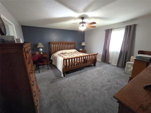 A bedroom with gray carpet, a wooden bed with a quilt, two nightstands with lamps, a ceiling fan with a light, dressers, and a window with gray curtains letting in natural light.