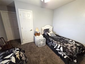 A bedroom with two twin beds covered in black and white floral bedding, a white nightstand with a lamp and decorative train, a fan on the floor, gray carpet, and pale gray walls. A wooden rocking chair sits in the corner.