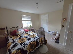 A bedroom with beige walls, an unshaded lightbulb on the ceiling, a bed with a sunflower-patterned comforter, a floor fan, boxes, and various items scattered around, with light coming through a window.