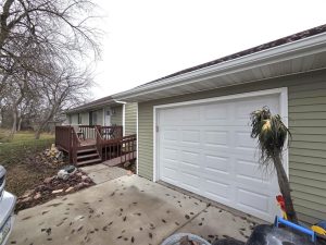 A green house with white trim and a white garage door, a wooden porch with steps, bare trees, and scattered leaves on the driveway. A tall plant stands near the garage.