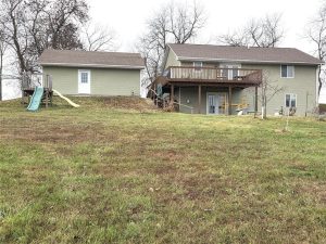 A grassy backyard with a two-story beige house featuring a raised wooden deck and stairs. To the left is a small outbuilding with a green slide attached. Leafless trees are in the background.