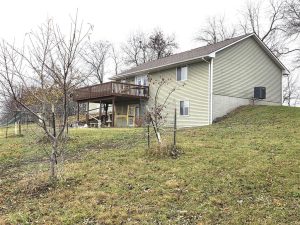A light green, two-story house with a raised wooden deck and stairs stands on a grassy slope. Leafless trees and a few young saplings are visible in the yard on a cloudy day.
