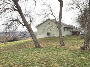 A single-story beige house with a wooden deck sits on a grassy, slightly sloped yard with two leafless trees in the foreground and a cloudy sky overhead.