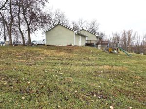A grassy backyard with a gentle slope, two light-green houses with decks, a parked car, bare trees, and a children's playset with a slide on the right side. The sky is overcast.