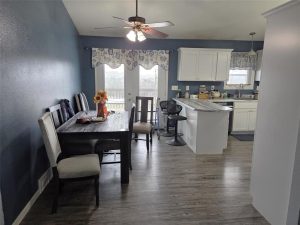 A cozy kitchen and dining area with blue walls, wood flooring, a ceiling fan, a dark wood dining table with four chairs, a kitchen island with two stools, white cabinets, and floral curtains on the windows and door.