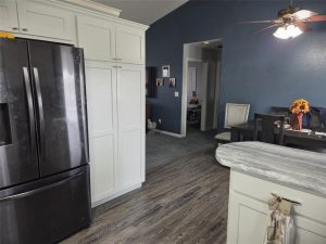 A kitchen with white cabinets, a black refrigerator, and gray wood floors. A dining area with dark table and chairs is visible, with a vase of sunflowers on the table. The walls are painted dark blue.