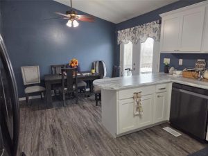 A kitchen with white cabinets and gray countertops is shown in the foreground, with a dining area featuring a dark table, mismatched chairs, and blue walls in the background. A ceiling fan and window with floral curtains are visible.