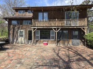 Two-story wooden house with a balcony and several large windows, sitting on a brick patio. A red cooler is on the ground near the center, and trees with bare branches surround the house.