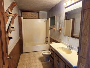A dated bathroom with a white bathtub/shower combo, toilet, long sink countertop, wooden cabinets, and wall hooks by the door. The upper wall and ceiling show signs of damage and exposed insulation.