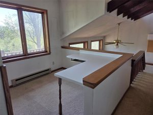 A small loft area with white walls and carpeting, featuring a built-in white desk under sloped ceilings, large windows with wooden trim, and a ceiling fan above a staircase. Trees are visible outside the windows.