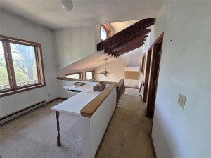 Loft area with white walls, slanted ceiling, exposed wooden beams, large windows, built-in desk, beige carpet, and a staircase with a wooden railing leading downstairs. Ceiling fan visible below.