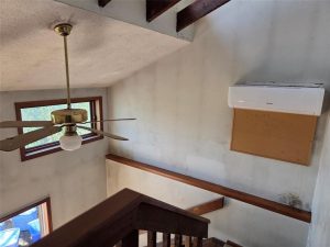A stairwell with wooden railings, a ceiling fan with a brass fixture, a wall-mounted air conditioning unit, and a corkboard on a white wall. Sunlight enters through two windows.