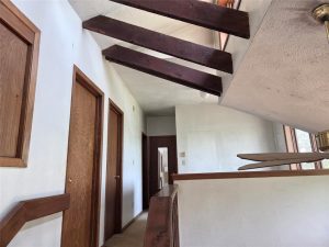 Interior hallway with white walls, wooden doors, and exposed wooden ceiling beams. A staircase with an open railing leads downward, and windows allow natural light into the space.