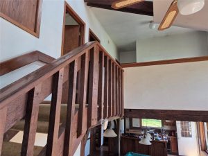 Interior view of a house with a wooden staircase and railings, beige carpeted steps, white walls, and ceiling fans. Downstairs, there is a kitchen area with hanging lights and visible windows.