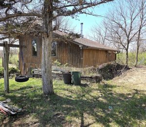 A rustic wooden house with a sloped roof sits among leafless trees. In the yard, there’s a tire swing, a wooden fence, garden tools, and trash bins on the grass under a clear sky.
