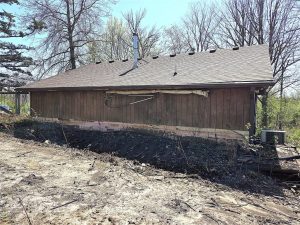 A weathered, one-story wooden building with a damaged exterior wall and missing roof shingles, surrounded by bare trees and overgrown vegetation on a patch of disturbed ground.