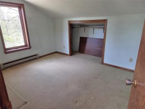 A small, empty room with beige carpet, white walls, a window with wooden trim, a baseboard heater, and an open closet with shelves and a large wooden panel inside.