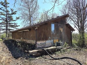A weathered, wooden house with a slanted roof sits among bare trees and overgrown vegetation. Exposed pipes, peeling paint, and debris are visible around the building on a sunny day.