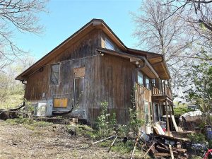 A weathered wooden house with peeling paint and boarded-up windows sits on uneven ground, surrounded by leafless trees and scattered debris under a clear blue sky.