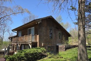A wooden house with a balcony sits on a grassy slope surrounded by trees and shrubs on a clear, sunny day. The home has large windows and an outdoor seating area below the deck.