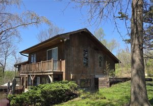 A wooden house with a balcony sits on a grassy slope surrounded by trees and shrubs on a clear, sunny day. The home has large windows and an outdoor seating area below the deck.